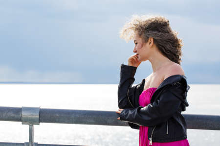 curly-haired Caucasian woman in a black leather jacket and a pink dress, stands on a bridge against...