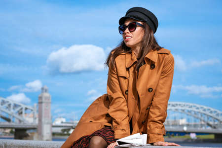 Young beautiful caucasian woman in a black cap and sunglasses in a coat on the embankment posing...