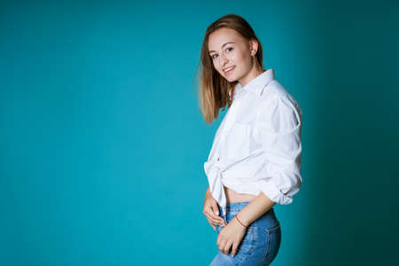 Young blond woman of caucasian ethnicity posing in a white shirt and jeans on a blue backgroundの写真素材