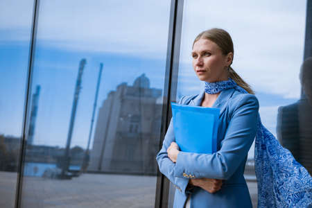 A business woman in a blue jacket holds a folder with papers in her hand in front of a glass office building. Concept of successful womenの写真素材