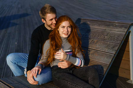 young couple of guy and girl with red hair of Caucasian appearance, in casual clothes, on a sunny day sitting in the park on a wooden bench in an embrace, happy relationship between a man and a womanの写真素材