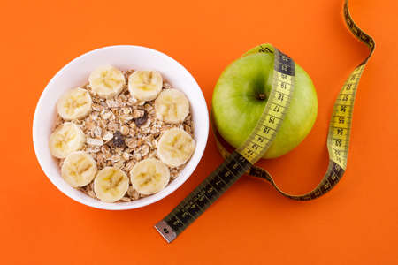 muesli with banana in a white plate on an orange background, green apple with a yellow measuring...