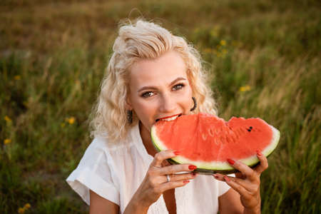 Close-up portrait of a woman biting a watermelon in nature. Beautiful girl of caucasian appearance eats a watermelon on a picnic.の写真素材