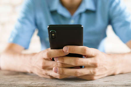 Young man holds phone in his hands and is texting. Guy sits at the table playing online games. Reads...