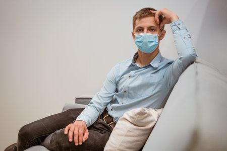 a young guy of Caucasian ethnicity in a shirt and a protective mask sits on the couch alone at home. Concept of being at home during global quarantineの写真素材