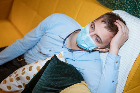 a young guy of European appearance, sitting comfortably on the couch in a medical mask. Concept to...