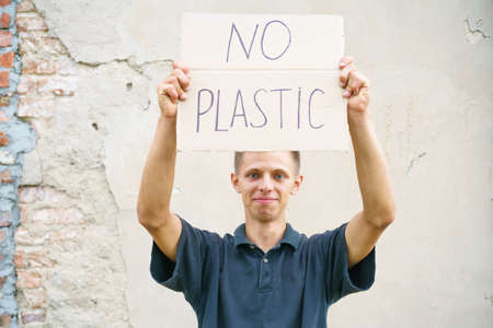 Caucasian guy holding the inscription no plastic. Environmental protection concept from garbage pollution . Young man protesting with placard banning plastic against cement wall backgroundの写真素材