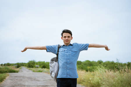 Happy young man posing with raised arms, standing on the road and looking into the distance. Happy guy in a blue shirt with a backpack. Free student enjoys vacationの写真素材