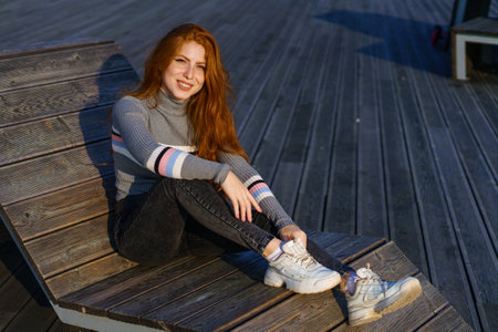 Happy young woman with long red hair of Caucasian nationality, is sitting in casual clothes in the park on a wooden deck on a sunny day and smilingの写真素材