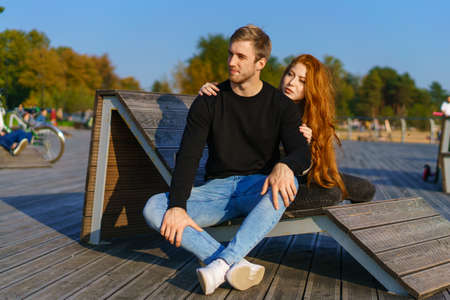 happy couple a guy and a girl with long red hair are sitting on a wooden deck in an embrace. Young man and woman of Caucasian ethnicity in casual clothes on a warm sunny day huggingの写真素材
