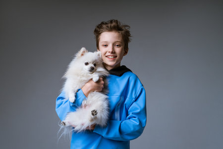 happy boy blond European appearance in blue clothes, holding a white dog on a gray background and smiling with snow-white teethの写真素材