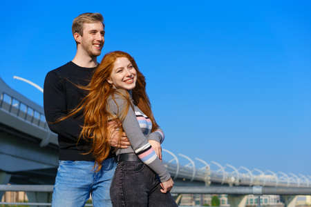 happy young couple man and woman with long red hair, stand against the background of a blue sky and...