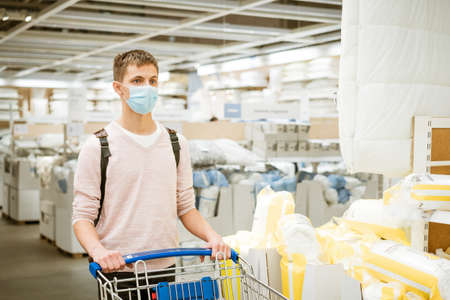 a guy of European appearance in a protective mask and casual clothes, with a trolley in the store in...