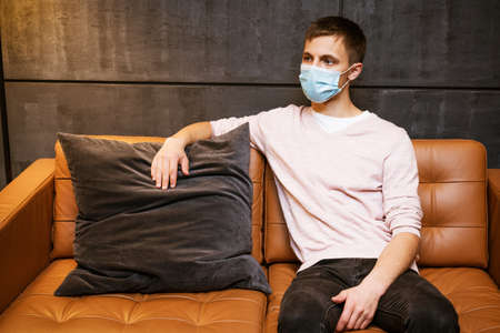 a young guy of Caucasian ethnicity in a shirt and a protective mask sits on the couch alone at home....