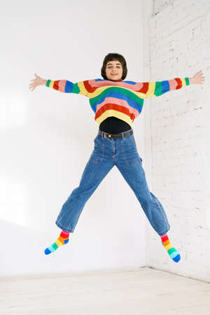 Happy girl in rainbow sweater jumps up on light background. Caucasian young woman jumping with...