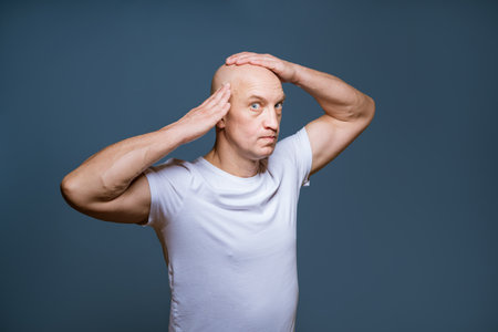 Funny bald man posing on a blue background with excited facial expressions and raised hands at his...