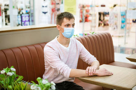 a young man of Caucasian appearance in casual clothes, sitting in a protective mask at a table in a cafe while waiting for an orderの写真素材