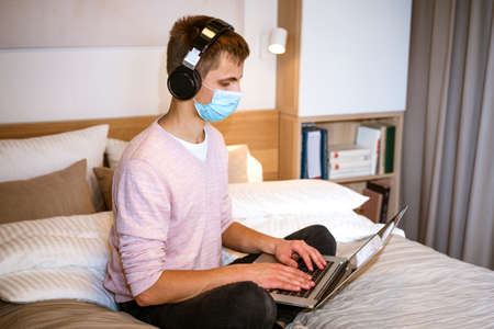 a young man of european appearance in casual clothes sits on a bed in a room at home wearing headphones and a protective mask at home with a laptop, the concept of remote work and educationの写真素材
