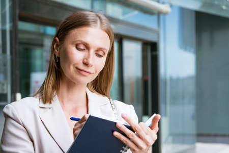Business woman writing with pen in notebook. Caucasian successful girl in light jacket stands near an office building. Recording a daily routineの写真素材