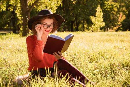 Woman in hat and red sweater and glasses reading a book in the park on a sunny autumn day