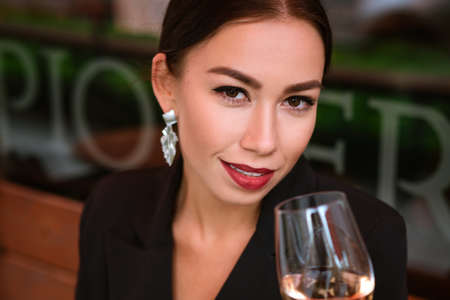 beautiful young woman of Caucasian ethnicity, beautiful makeup and red lipstick on her lips. In black jacket and earrings, he sits in street cafe with glass of wine and smiles with snow-white teeth.の写真素材