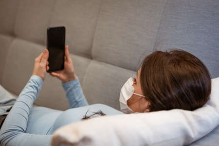 tired caucasian woman in casual clothes in a protective mask on the couch with a phoneの写真素材