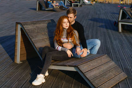 young couple of guy and girl with red hair of Caucasian appearance, in casual clothes, on a sunny day sitting in the park on a wooden bench in an embrace, happy relationship between a man and a womanの写真素材