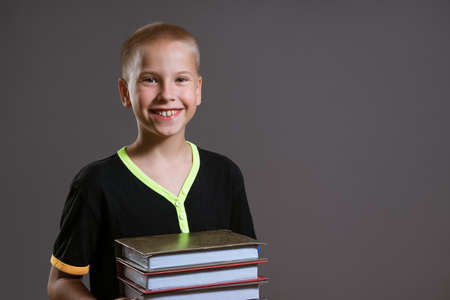Cheerful caucasian boy in a black t-shirt holds a stack of books on a gray background. Education conceptの写真素材