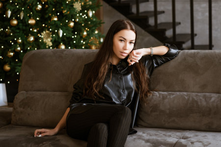 portrait of a beautiful young adult brunette woman sitting on the sofa in the living room. the girl is wearing a black leather jacket and tight dark trousers. the model looks at the camera and smiles.の写真素材