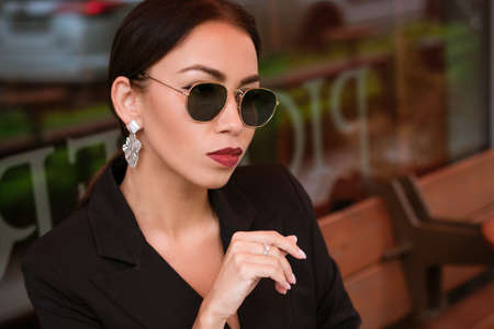 Portrait of a young business woman, brunette Caucasian appearance in a black suit and an earring sits in sunglasses in a cafe at a table. Red lipstick on lips. A business meetingの写真素材