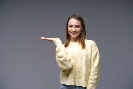 Portrait of a young woman of Caucasian ethnicity shows with her hand to the side in a yellow sweater on a gray background, the concept of advertising goods on her handの写真素材