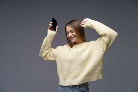 Young cheerful woman of Caucasian ethnicity in a yellow sweater on a gray background is dancing with a phone in her hand. Happy girl listening to music on the phone dancing raising her hands upの写真素材