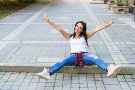 Portrait of young beautiful brunette woman with long hair in blue jeans and white t-shirt sitting on sidewalk on city street. Lifestyle cheerfully spread her arms to the sides. Happy caucasian womanの写真素材