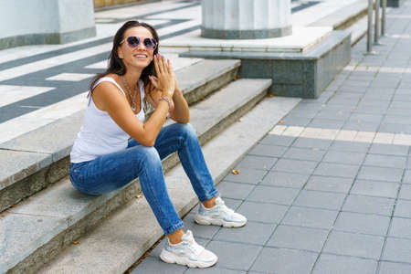 Portrait of beautiful woman in casual clothes and sunglasses sitting on stairs in the city. Stylish adult girl smiling cute.の写真素材