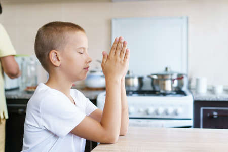 Caucasian boy prays at home at the table before eating. Stay home and pray to god. Online church service on sunday. Childs hand folded in prayer. Son sits in the kitchen and prays to godの写真素材