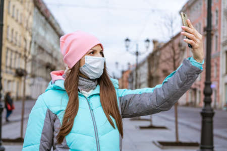 Young woman of Caucasian ethnicity in a warm jacket, in a protective mask, takes pictures on the...