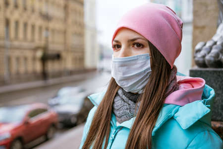 a cute young woman of Caucasian ethnicity, in a warm jacket and a pink hat, in a medical mask, walks...