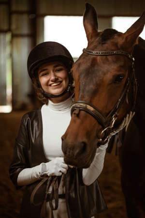 Young woman rider with a brown beautiful horse, standing nearby in jockey clothes, selective focus....
