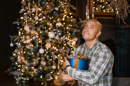 A Caucasian cheerful young handsome man in a good mood poses with a wrapped gift near a decorated...