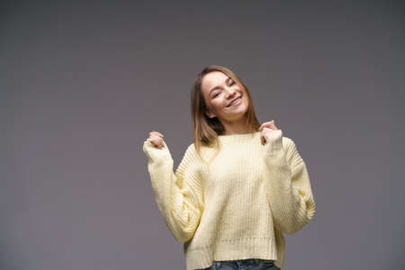 Portrait of a beautiful young girl of Caucasian ethnicity in a yellow sweater on a gray background cute smiling. Keeps his hands to his face.の写真素材