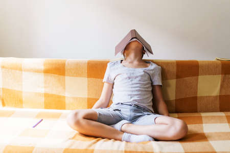 Caucasian boy sitting on couch covered his head with a notebook from fatigue to solve homework. A guy in casual clothes takes a break between lessons sits in meditationの写真素材
