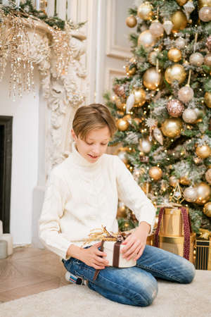 Cute boy is sitting by the christmas tree with gifts. Caucasian teenager in warm knitted light sweater sits on the floor of house with festive new years decor in the room. Holding gift in his handの写真素材
