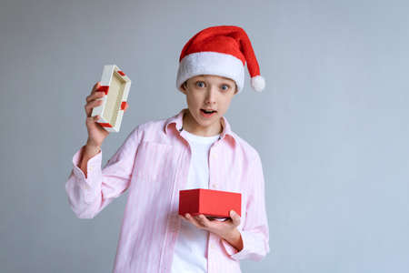 Caucasian guy in santa claus hat and shirt opens gift and looks with surprise on a light background in the studio. Happy boy received a box with a giftの写真素材
