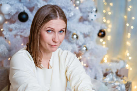 Portrait of caucasian woman in light light jumper sitting by christmas tree with a pensive look. Festive decor behind the back, the garland is litの写真素材