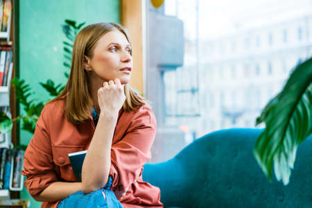Beautiful caucasian girl spends her free time at home, sitting on couch, reading a book and looking...