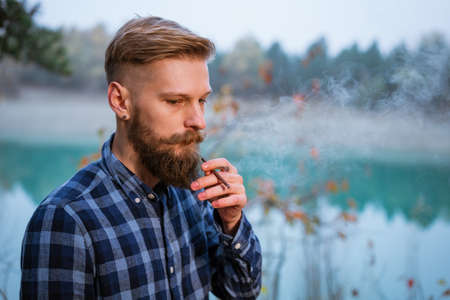 The bearded man smokes. Handsome stylish man in plaid shirt with cigarette. Nicotine addiction and bad habits. Caucasian guy stands in the evening by the lake in the forestの写真素材