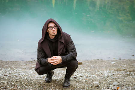 Bearded man in casual clothes with hood and glasses stands on shore of turquoise lake and looks to side. Relaxation and solitude with nature in autumn. Caucasian cute kidの写真素材