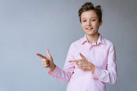 A cute boy of European appearance, a blondie in a pink shirt , points to the side with his fingers on a light background and smiles with snow-white teethの写真素材