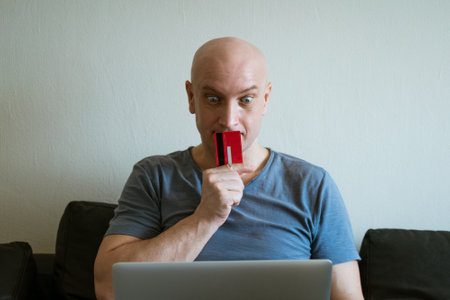 an adult bald man in a blue T-shirt is sitting at home on the couch with a bank card in his hand at...