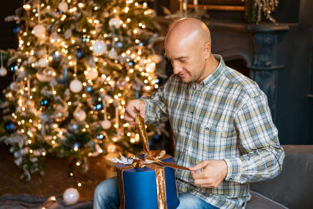 Happy bald man in a shirt opens a Christmas present while sitting on the sofa against the background of a Christmas treeの写真素材